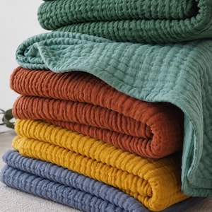 Puede incluir: Una pila de cinco mantas dobladas con un tejido de textura de gofre. Las mantas están dispuestas en colores verde oscuro, verde salvia, naranja quemado, amarillo mostaza y azul pizarra, mostrando un patrón de tela suave y cuadriculado.