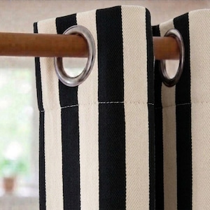 Op de afbeelding: Een gordijnpaneel met een verticaal zwart en gebroken wit visgraatpatroon. De stof hangt aan een houten roede door middel van twee grote, ronde, zilverkleurige metalen ringen die gelijkmatig over de bovenste zoom zijn verdeeld.