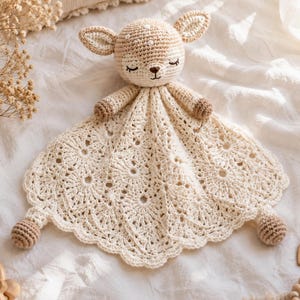 May include: A crochet security blanket featuring a stuffed deer head with closed eyes and small ears. The blanket body is made of cream-coloured yarn with a detailed, openwork floral pattern and scalloped edges, finished with small brown crochet spheres.