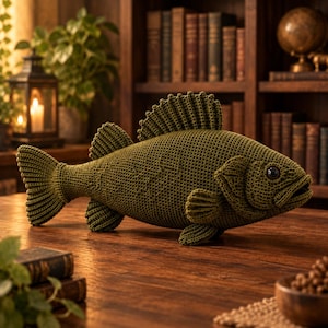 May include: A stuffed fish toy crafted from olive green yarn using a crochet technique. The fish features a prominent dorsal fin, a detailed tail, side fins, and a single dark eye. The texture is consistent throughout the body, showcasing the intricate loops of the crochet work in a solid, muted green colour.