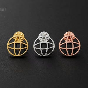 Puede incluir: Tres pines circulares de solapa con un diseño de globo estilizado y líneas que se cruzan. Los pines tienen acabados en tonos oro, plata y oro rosa. Cada pin incluye un cierre metálico. El texto OMNIVERSE PROPS es visible en la esquina.