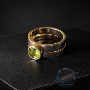 May include: A two-tone ring featuring a round, faceted lime green gemstone set in a gold bezel. The ring band is constructed from a wide silver-colored metal base topped with a thinner gold-colored band, creating a stacked appearance.
