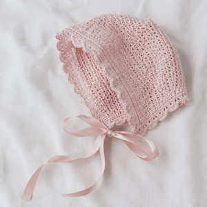 May include: A light pink crochet bonnet featuring a delicate openwork stitch pattern and a scalloped edge trim. The headwear is secured with a matching satin ribbon tie that is tied into a bow at the front, providing a soft and textured appearance.