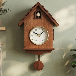 May include: A wooden cuckoo clock shaped like a house with a gabled roof, a white circular clock face with black numerals and hands, a small bird figure in an upper window, and a round wooden pendulum hanging from the base of the clock body.