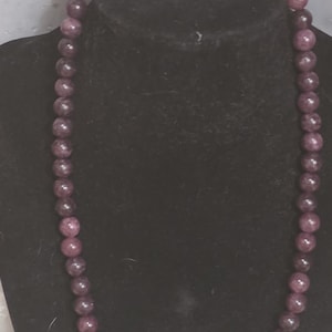 May include: A beaded necklace featuring a continuous strand of smooth, dark purple spherical beads. The necklace includes two tan, textured accent beads near the gold-toned lobster clasp and a central arrangement of two tan textured beads flanking a small, smooth, light brown heart-shaped bead.
