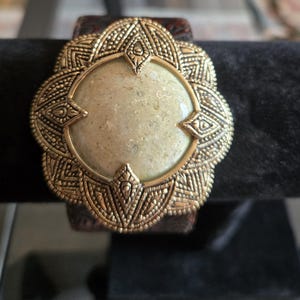 Peut inclure: Un bracelet manchette large composé d'une bande en cuir marron foncé avec un motif gaufré. La pièce centrale est une grande pierre beige ronde et texturée, sertie dans un cadre en métal doré vieilli aux détails complexes et festonnés.
