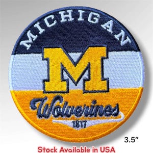 May include: A circular embroidered patch featuring a navy blue top section with the word MICHIGAN in white, a white middle section with a large yellow letter M, and a yellow bottom section with the word Wolverines in navy blue and 1817 below it.