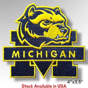 May include: An embroidered patch featuring a navy blue and yellow wolverine head logo positioned above a horizontal bar with the text MICHIGAN in yellow block letters, all set against a large navy blue letter M, measuring 4 inches by 3.5 inches.