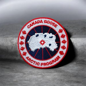 May include: A circular embroidered patch featuring a white map of the Arctic on a dark blue background with red radial lines. The white border contains the red text CANADA GOOSE and ARCTIC PROGRAM, separated by small red maple leaf icons.