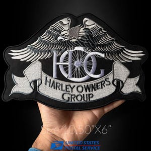 May include: A black embroidered patch featuring a silver eagle with spread wings above a motorcycle wheel and the text HOG HARLEY OWNERS GROUP. The patch measures 10.50 inches by 6 inches, with a detailed silver thread design on a black fabric base.