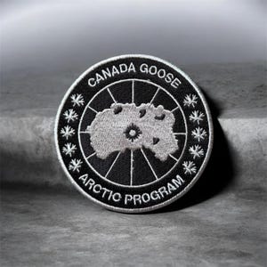 May include: A circular embroidered patch featuring a black background with white text reading CANADA GOOSE and ARCTIC PROGRAM. The center displays a light gray map graphic with a small star icon, surrounded by white maple leaf symbols.