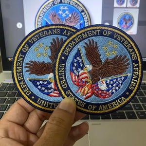 May include: Two circular embroidered patches featuring the Department of Veterans Affairs seal. Each patch displays a brown eagle clutching a flag over a light blue background, surrounded by a dark blue border with gold text reading Department of Veterans Affairs United States of America.