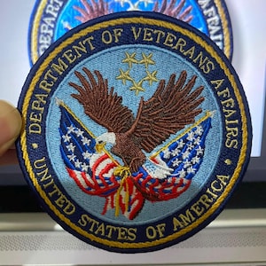 May include: A circular embroidered patch featuring the Department of Veterans Affairs seal. It displays a brown eagle with wings spread over two crossed American flags on a light blue background, surrounded by a dark blue border with gold text.