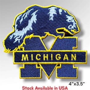 May include: An embroidered patch featuring a dark blue wolverine silhouette with light blue highlights positioned above a large dark blue letter M. A horizontal dark blue bar across the M contains the yellow text MICHIGAN. Measures 4 inches by 3.5 inches.