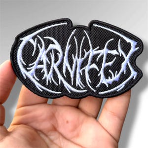 May include: A black embroidered patch with a white, stylized, branch-like font that reads Carnifex. The patch has a rounded, irregular shape with a thick black border and a textured fabric surface, featuring intricate white thread detailing.