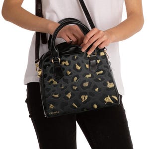 A person wearing a white t-shirt and black pants holds the handles of a structured black and grey leopard print handbag with gold details.