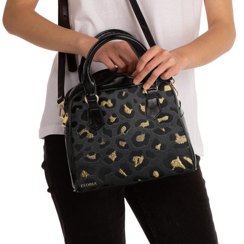 A person wearing a white t-shirt and black pants holds the handles of a structured black and grey leopard print handbag with gold details.