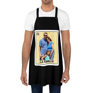 May include: Black apron featuring a family photo in a rectangular frame. The photo shows a family of three with the text "LA FAMILIA" below. The apron has black straps and a button closure.