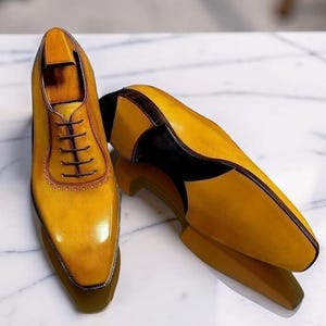 May include: A pair of mustard yellow oxford shoes with dark brown accents and dark brown laces. The shoes feature a polished finish, a pointed toe, and a wooden shoe tree inserted into the upright shoe. The design includes decorative stitching.