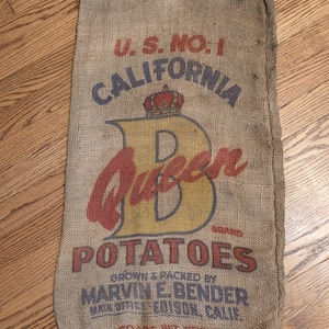 May include: A rectangular burlap sack featuring the printed text U.S. NO. 1 CALIFORNIA QUEEN BRAND POTATOES GROWN & PACKED BY MARVIN E. BENDER MAIN OFFICE EDISON, CALIF. 60 LBS. NET WEIGHT. The design includes a large yellow letter B and a red crown.