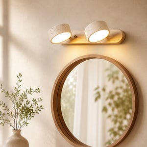 May include: A round wooden-framed mirror mounted on a wall below a horizontal light fixture. The light fixture features a light-colored wooden base with two cylindrical, speckled, off-white spotlights that emit a warm, downward-facing glow.