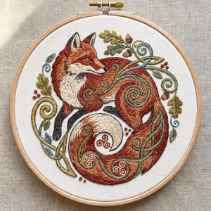 Könnte beinhalten: Ein runder Holzstickrahmen mit einer detaillierten Stickerei eines zusammengerollten Rotfuchses. Der Fuchs ist umgeben von gestickten grünen Eichenblättern, braunen Eicheln und verschlungenen Rankenmustern mit kleinen roten Beeren.