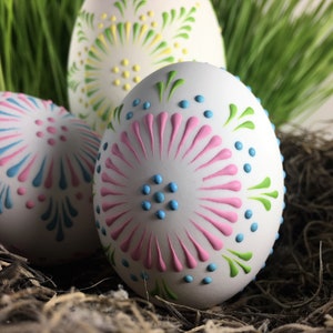 Pode incluir: Três ovos de Páscoa brancos decorados com desenhos de tinta de diferentes cores. Os ovos estão em um ninho de palha marrom.