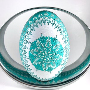 May include: A white egg with a teal and white intricate design. The egg is decorated with small white dots and is sitting on a silver stand.