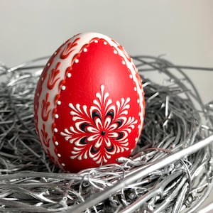 May include: A decorative red Easter egg with white and black floral designs, resting in a silver nest. The egg features intricate patterns and a glossy finish, perfect for Easter decor.