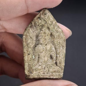 May include: A small, pointed, beige-colored clay amulet featuring a relief carving of a seated figure in a meditative pose. The surface has a rough, weathered texture with visible dark speckles and intricate embossed patterns surrounding the central figure.