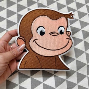 May include: A brown and tan embroidered patch featuring the head and shoulders of a cartoon monkey with large black eyes, a wide smile, and a small tuft of hair on top, finished with a thick white border around the entire character shape.