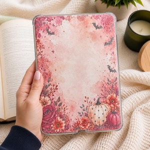 Puede incluir: Una funda rectangular para tableta con un estampado estilo acuarela que muestra un arreglo floral en rosa y rojo, calabazas con lunares y pequeñas siluetas de murciélagos negros sobre un fondo rosa claro con estrellas.