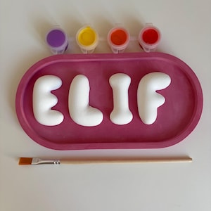 May include: A pink oval tray containing four white letters spelling ELIF, accompanied by a strip of four small paint pots in purple, yellow, orange, and red, and a single wooden paintbrush with a flat tip for a craft activity set.