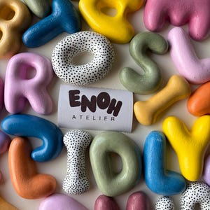 Pode incluir: Uma coleção de letras e formas tridimensionais arredondadas e coloridas em tons de rosa, amarelo, azul, laranja e verde sálvia, junto com peças de bolinhas pretas e brancas, cercando uma etiqueta retangular branca com o texto ENOH ATELIER.