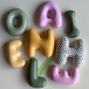 Peut inclure: Un ensemble de neuf lettres et formes en céramique à la finition brillante, déclinées en vert sauge, rose pastel, jaune moutarde et une pièce blanche à pois noirs. Une petite étiquette verte portant le texte @Enshatelier est présente.