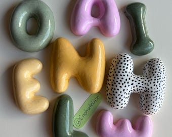 Puede incluir: Una colección de nueve letras y formas de cerámica en varios colores, incluyendo verde salvia, rosa pastel, amarillo y una pieza blanca con motas negras. Las letras son redondeadas y brillantes, con una etiqueta verde que dice @Enshatelier.