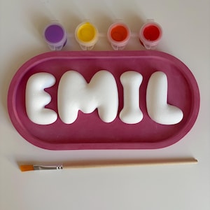 Puede incluir: Una bandeja ovalada rosa que contiene letras 3D blancas que forman la palabra EMIL, acompañada de cuatro pequeños botes de pintura en morado, amarillo, naranja y rojo, y un pincel de madera con cerdas naranjas debajo.