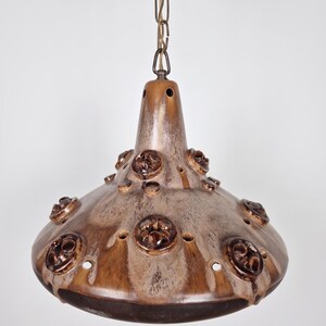 Peut inclure: Une suspension en céramique en forme de cloche avec une finition émaillée coulante brune et crème. La surface est décorée de médaillons floraux circulaires en relief et de petites découpes circulaires, suspendue par une chaîne en métal.