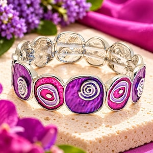 May include: A stretch bracelet featuring alternating silver-tone metal links and circular enamel plaques. The plaques display a vibrant colour palette of deep purple, magenta, and light lavender, each accented with a raised silver spiral design.