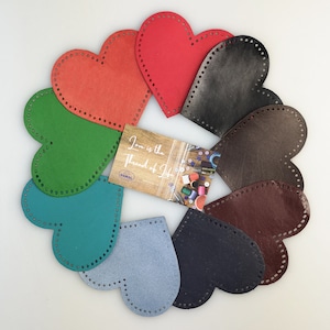 Puede incluir: Diez formas de corazón de cuero en varios colores, incluyendo rojo, verde, azul, marrón y negro. Los corazones tienen agujeros alrededor de los bordes y están dispuestos en un círculo. Una pequeña tarjeta con el texto "El amor es el hilo de la vida" está en el centro del círculo.