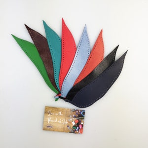 May include: Seven leather bookmarks in a variety of colours, including green, brown, teal, red, blue, black, and navy. The bookmarks are shaped like leaves and have a stitched edge. The bookmarks are arranged in a fan shape. A small card with the text "Love is the Thread of Life" is in the foreground.