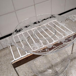 Puede incluir: Un plato de servicio de vidrio transparente con un patrón floral grabado y dos asas, apoyado sobre una rejilla de alambre blanca. Una regla de madera está colocada debajo del plato, mostrando una medida de unos 28 cm de ancho.