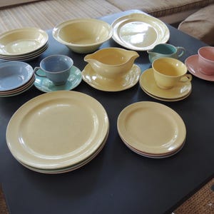Peut inclure: Un ensemble de vaisselle en céramique comprenant des assiettes plates, des assiettes à salade, des bols et une saucière jaunes. Le service inclut des tasses et soucoupes bleu pastel, rose et sarcelle, ainsi qu'un petit pot à lait sarcelle.