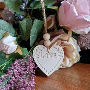 May include: A white heart-shaped ornament featuring the word LOVE embossed in the centre, accented with leaf motifs and small dots. The ornament hangs from a jute twine string adorned with two small, round, light-coloured wooden beads.
