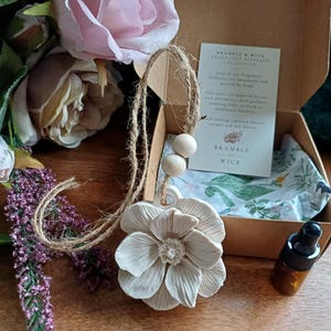 May include: A white ceramic flower ornament hanging from a jute twine string with two wooden beads, placed next to an open cardboard box containing a small amber glass dropper bottle and a white card with the text Bramble and Wick.
