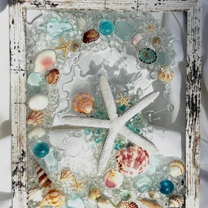 May include: A rectangular wall art piece in a distressed white wooden frame featuring a clear resin base embedded with various seashells, small starfish, and smooth blue and clear glass pebbles, centered with a large white starfish.