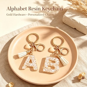 Può includere: Due portachiavi in resina a forma di lettere A e B, riempiti con frammenti di pietra bianchi e beige. Ogni portachiavi include componenti dorati, un piccolo ciondolo a cuore e una nappa bianca. Testo: Alphabet Resin Keychain.