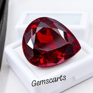May include: A pear-shaped, deep red faceted gemstone resting in a white display box. The stone features intricate geometric cuts that reflect light. To the left, a pair of black metal tweezers is visible. The text Gemscarts is printed in black.