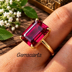 Op de afbeelding: Een gouden ring met een grote, rechthoekige, gefacetteerde dieprode edelsteen die door vier klauwen wordt vastgehouden. Het woord Gemscarts staat in zwarte letters op de huid bij de ring. De ringband is van glad, gepolijst goud en toont de rode steen.