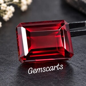 May include: A deep red, emerald-cut gemstone with sharp, clean facets that reflect light. The stone is held by metal tweezers and features the text Gemscarts printed in white below it, showcasing a vibrant, saturated crimson color throughout.